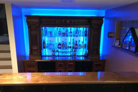 A stunning home bar setup, perfect for entertaining. This image shows how to build a bar with a wooden countertop, a backlit liquor cabinet filled with bottles, and vibrant blue ambient lighting.