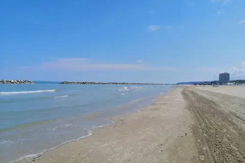 Spiaggia del lido di Fermo con mare calmo, frangiflutti in pietra e un palazzo sullo sfondo.