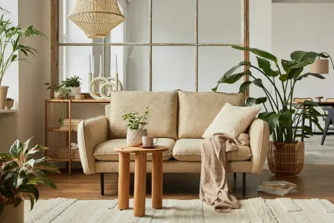 Przytulny salon w stylu cottage: beżowa kanapa, drewniany stolik, dużo roślin i pleciony klosz lampy tworzą ciepłą atmosferę.