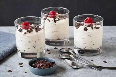Drei Gläser mit cremigem Dessert, garniert mit Himbeeren und Schokoladenstückchen. Ein Schälchen mit Schokostückchen und Löffel daneben. Ein leckeres Herrencreme Rezept zum Nachmachen.