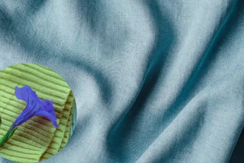 A blue tencel fabric drapes softly, hinting at its smooth, natural feel. A purple flower rests on a textured yellow surface.