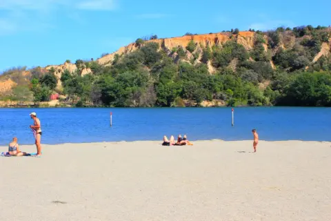 Plage de sable fin au lac de Vinça, avec des baigneurs profitant du soleil et de la vue sur la colline verdoyante.