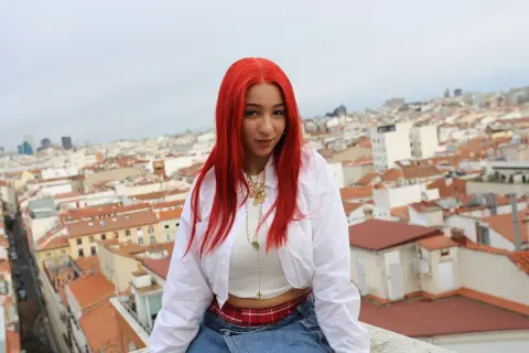 Joven con cabello rojo vibrante, como una rosa encendida, posa sobre tejados urbanos.