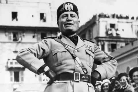 Benito Mussolini, con uniforme militar y gorra, sonríe ante una multitud. Su ascenso al poder es un capítulo oscuro de la historia.