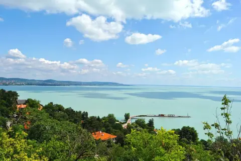Widok na turkusowe jezioro Balaton z zielonymi wzgórzami i domami. Na wodzie widać molo i żaglówki.