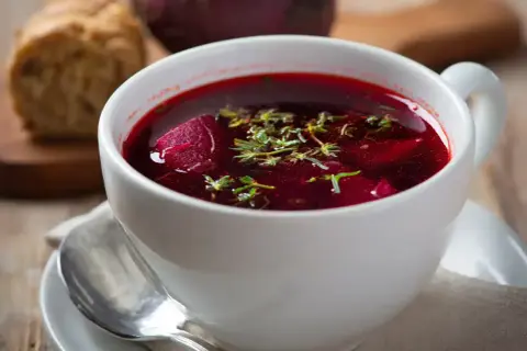 Aromaticzny barszcz czerwony domowy, z kawałkami buraka i ziołami, podany w białej filiżance z łyżeczką.