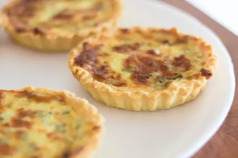 Trois petites quiches sans œuf dorées sur une assiette blanche. La pâte est croustillante et la garniture semble crémeuse.