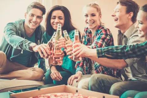 Grupa przyjaciół wznosi toast butelkami piwa, śmiejąc się i jedząc pizzę. Nawet trudne pytania nie psują im nastroju.