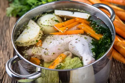 Pentola con brodo di pollo ricetta della nonna: pollo, carote, sedano, cipolla e prezzemolo in cottura su un tavolo di legno.
