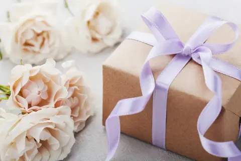 Un cadeau d'anniversaire fait maison, joliment emballé dans du papier kraft et un ruban lilas, entouré de roses pâles.