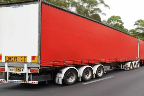 Un long camion 60 tonnes, avec une remorque rouge vif, roule sur une route bordée d'arbres.