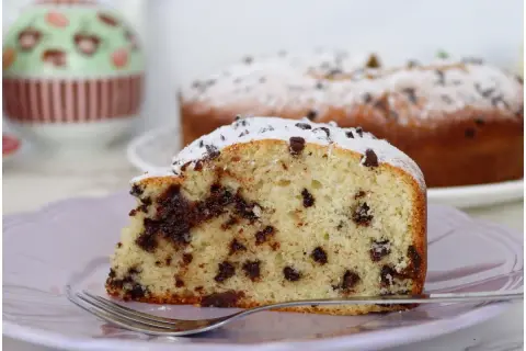 Una fetta di torta soffice con tante gocce di cioccolato, cosparsa di zucchero a velo, su un piatto lilla con una forchetta.