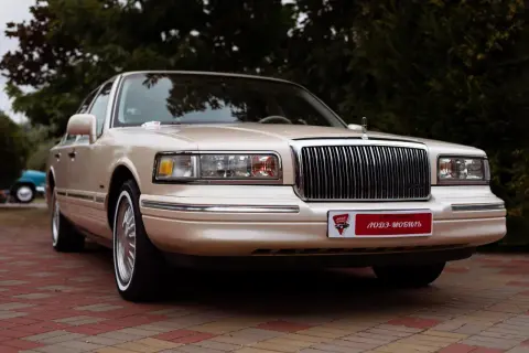 Beżowy Lincoln Town Car, przyszły youngtimer, lśni na tle zieleni. Klasyczna elegancja, która wciąż zachwyca.
