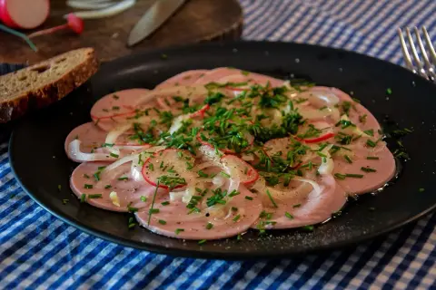 Saure Wurst mit Zwiebeln, Radieschen und Schnittlauch auf schwarzem Teller, dazu Brot.