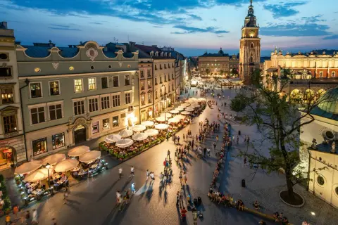 Wieczorny gwar na Rynku Głównym w Krakowie. Ludzie spacerują, siedzą w ogródkach kawiarnianych. W tle Sukiennice i wieża ratuszowa. To serce miasta, jego **sarbinowo centrum**.