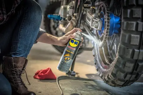 Persona aplica cera en aerosol WD-40 para limpiar la cadena de la moto, preparándola para la próxima aventura.