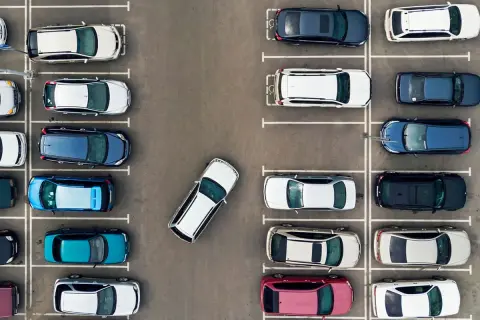 Widok z góry na parking z wieloma samochodami zaparkowanymi prostopadle. Jeden biały samochód jest zaparkowany pod kątem.