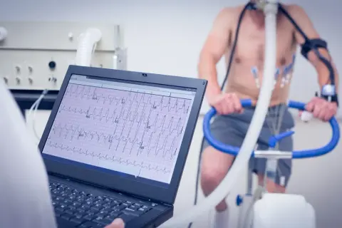 Un homme subit un test d'effort cardiologue sur un vélo d'appartement, avec un électrocardiogramme affiché sur un écran d'ordinateur.