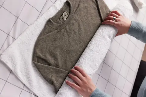Hände rollen einen braunen Kaschmirpullover auf einem weißen Handtuch auf, um ihn nach dem Kaschmir waschen zu trocknen.