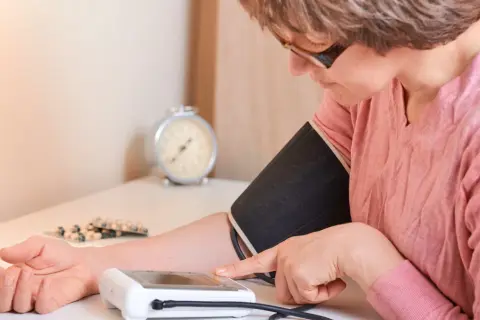 Kobieta mierzy ciśnienie, zmagając się z **wysokim ciśnieniem**. Na stole leżą leki.