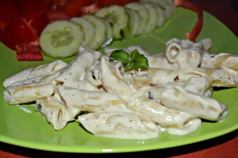 Penne w kremowym sosie z mozzarellą, udekorowane bazylią. Obok plasterki ogórka i papryka.
