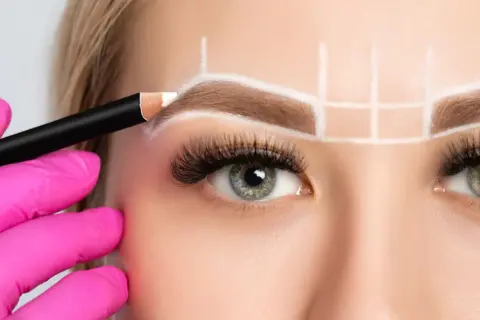 Manos enguantadas en rosa dibujan con lápiz blanco para crear cejas perfectas, marcando la simetría y el diseño.