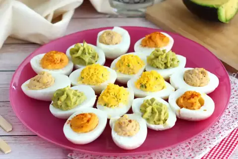 Assortiment d'œufs mimosa colorés sur une assiette rose. Des garnitures vertes, jaunes et orangées apportent une touche festive.