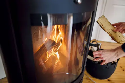 Hände legen Holzscheite in einen modernen Kamin, der brennt. Informieren Sie sich, welche Kamine noch erlaubt sind.