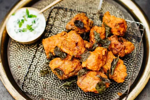 Knusprige chicken pakora, garniert mit frischen Kräutern, auf einem Gitterrost serviert. Daneben eine Schale mit Joghurt-Dip.
