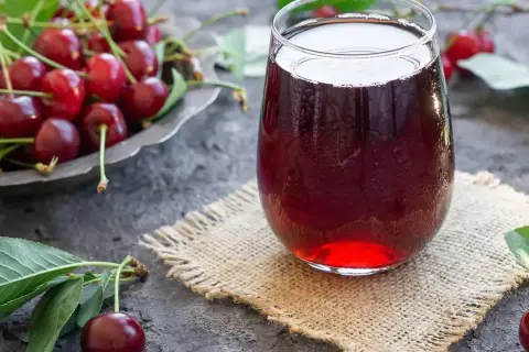 Szklanka orzeźwiającego soku z wiśni, obok miseczki pełnej świeżych owoców. Idealny na lato, prosty sok z wiśni przepis.
