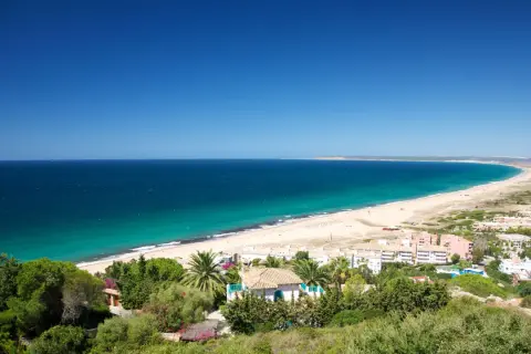 Vista panorámica del pueblo y paseo marítimo de Zahara de los Atunes, con su extensa playa de arena dorada y el mar azul turquesa.