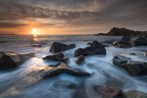 Available light photos capture a serene sunset over a rocky coastline. Water flows gently around dark, wet stones, reflecting the warm hues of the sky.