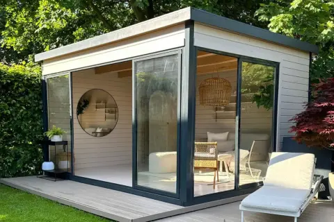 Modernes Gartenhaus mit Glasfront, das zum Streichen einlädt. Gemütliche Inneneinrichtung mit Rattankorb-Lampe und Liegestuhl draußen.