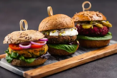 Drei appetitliche vegetarische Gerichte: Burger mit bunten Patties, frischem Gemüse und cremigen Saucen auf rustikalem Holzbrett.