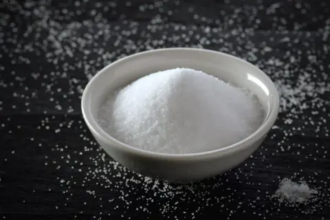 Biała miseczka pełna drobnego, krystalicznego proszku, który może być słodzikiem, np. maltitolem.