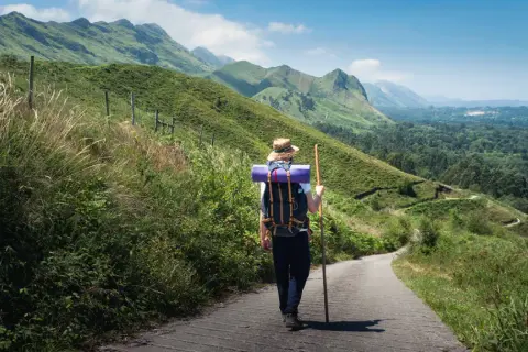 Pielgrzym w kapeluszu i z plecakiem idzie ścieżką przez zielone wzgórza. To jedna z malowniczych tras Camino de Santiago.