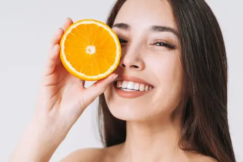 Lächelnde Frau hält Orangenscheibe vor ihr Auge. Vitamin C kann bei Rosacea helfen, die Haut zu beruhigen.