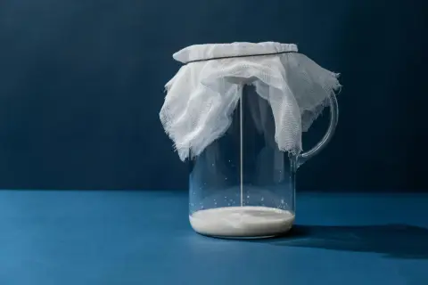 Un pichet en verre contient du kéfir, recouvert d'une étamine blanche retenue par un élastique. La boisson fermentée coule doucement.
