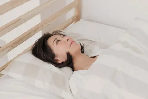 A woman lies in bed, looking up thoughtfully. Is it safe to sleep while dehydrated? Her expression suggests she's pondering this question.