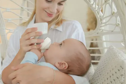 A mother smiles lovingly while holding her baby for upright bottle feeding. The baby is nestled in her arms, taking milk from the bottle.