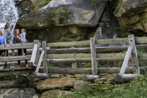 Famiglia felice sul sentiero delle cascate, con una cascata fragorosa alle spalle e un ponte di legno rustico.