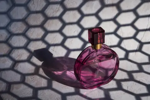 Różowa buteleczka perfum Dior J'adore, oświetlona słońcem, rzuca cień o geometrycznym wzorze plastra miodu.