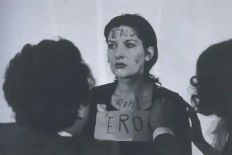 Marina Abramović w performansie "Rhythm 0" z napisem "END" na czole i "WOMAN ERO" na klatce piersiowej.