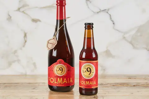 Due bottiglie di birra ambrata "La 9" del Birrificio L'Olmaia, una da 750ml e una più piccola, su un tavolo di legno.
