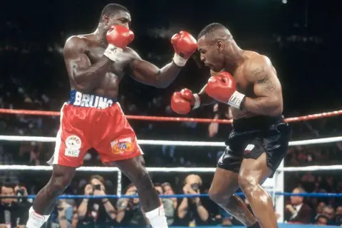 Two boxers, one in red shorts with "BRUNO" on the waistband, the other in black shorts, face off in a tense boxing match. The pressure fighter in red throws a punch.