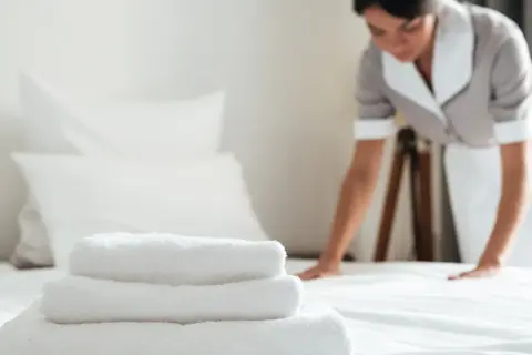 Pokojówka w hotelu dba o czystość i porządek. Na pierwszym planie stos białych ręczników, w tle kobieta poprawia pościel.
