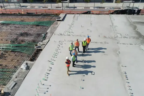 Grupa budowlańców na placu budowy, dyskutująca o tym, ile kosztuje kubik betonu. Widać zbrojenie i świeżo wylany beton.