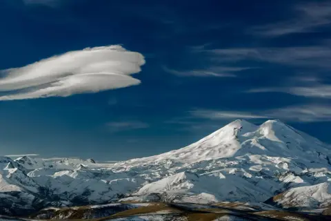 Pokryty śniegiem najwyższy szczyt Kaukazu, Elbrus, góruje nad ośnieżonymi pasmami górskimi pod błękitnym niebem z charakterystyczną chmurą.