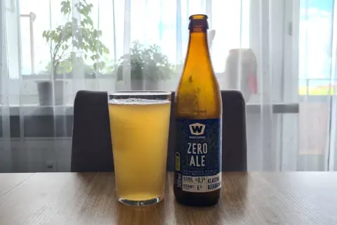 Szklanka i butelka piwa bezalkoholowego "Zero Ale" na drewnianym stole. Etykieta informuje, co zawiera piwo bezalkoholowe: ekstrakt 6%, alkohol ≤0.5%.