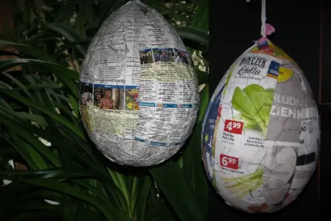 Jak zrobic jajko z gipsu w balonie? Dwa papierowe jajka, jedno z gazet, drugie z reklam, wiszą na tle zielonych liści.